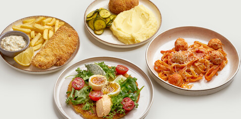 A variety of delicious and appetizing dishes are arranged on a white surface. The dishes include a bowl of creamy soup, a plate of meatballs and pasta, a plate of mashed potatoes