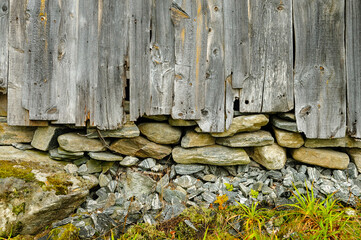 A rustic wooden structure stands with its aged planks weathered by time, resting on a foundation of smooth stones and surrounded by vibrant greenery as sunlight delicately illuminates the scene.