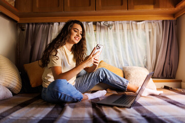 Woman on the bed of her camper van while using mobile with her laptop. Digital nomad