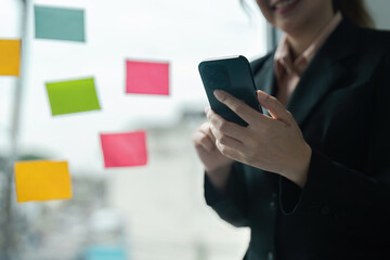 Professional Businesswoman Using Smartphone in Modern Office with Colorful Sticky Notes on Glass Wall