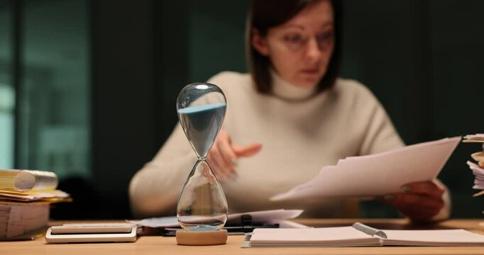 Woman looks through documents with an hourglass ticking away the time late at night in the office