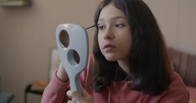 Cute teenage girl putting on make-up using mascara looking at mirror sitting on bed at home. Beauty routine and cosmetics concept.