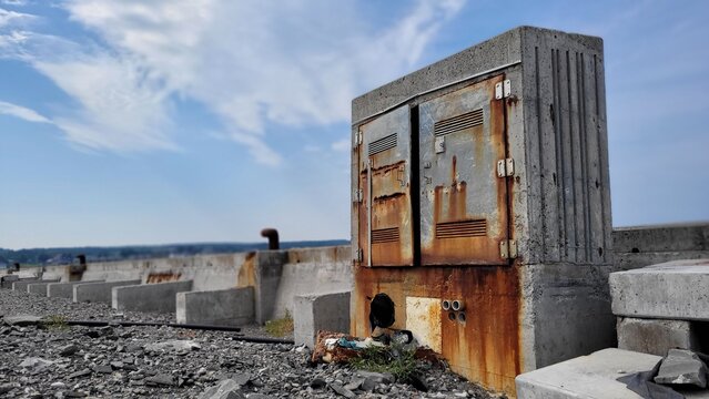 Vieux cabinet &eacute;lectrique rouill&eacute; abandonn&eacute; cotoyant une bordure de ciment surmont&eacute; de bornes d'amarrage a bateaux sur les vestiges d'un vieux quai. sous un ciel bleu parsem&eacute; de nuages.