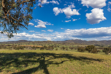 A vast rural landscape unfolds with gentle rolling hills beneath a vibrant blue sky dotted with...