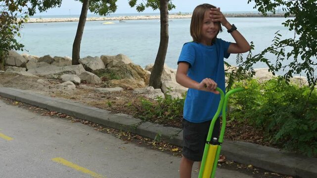 A boy in a blue shirt enjoys hopping on a pogo stick by the seaside, with trees and rocks in the background. The calm sea and distant pier add a peaceful vibe