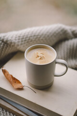 A warm cup of coffee rests on a stack of books, surrounded by soft blankets and autumn leaves, creating a tranquil atmosphere by the window.