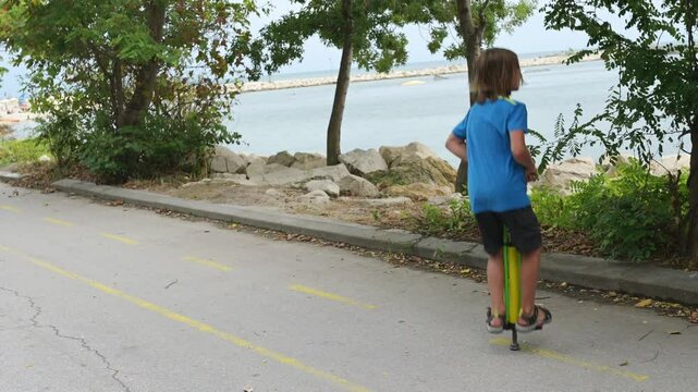 A young boy on a green pogo stick plays joyfully by the seaside, near rocks and trees. The tranquil ocean and a distant pier create a serene and adventurous atmosphere