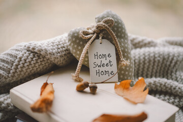 A heart-shaped decorative piece with a tag that says Home Sweet Home is placed on a knitted blanket, surrounded by fallen autumn leaves, creating a warm, inviting atmosphere.