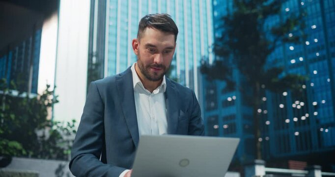 Portrait of a Cheerful Businessman in a Blue Suit Sitting in a Park in a Modern Urban Plaza, Typing on His Laptop Computer. Manager is Responding to Urgent Emails and Managing His Project Deadlines