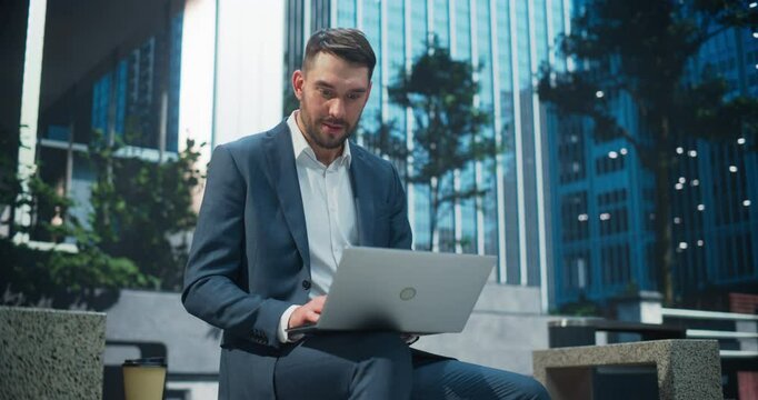 Portrait of an Excited Businessman in a Suit Sitting on a Bench in a Busy City Square, Typing on His Laptop Computer. Caucasian Man Is Finalizing a Presentation for an Important Client Meeting