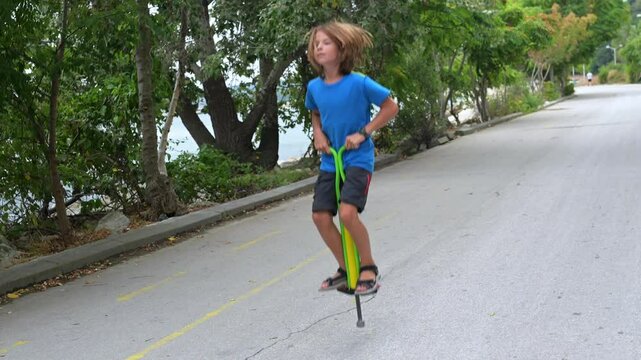 A boy in a blue shirt enjoys hopping on a pogo stick by the seaside, with trees and rocks in the background. The calm sea and distant pier add a peaceful vibe