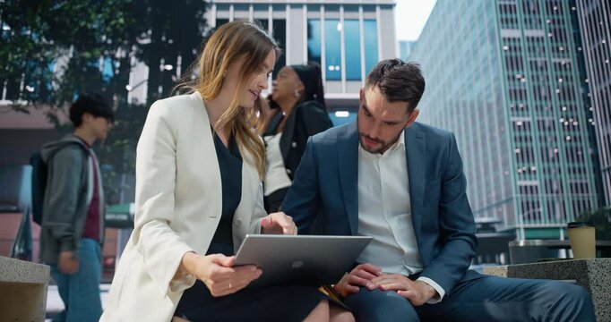 Female Team Leader and Male Manager Sit Side by Side on a Park Bench in a Green Urban Space, Both Focused on Data on Laptop Computer. They Are Editing the Final Draft of a Strategic Business Plan