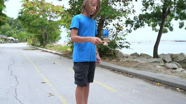 A young boy in a blue shirt plays with a kendama toy by the seaside, surrounded by trees and rocks. The calm water and peaceful setting enhance the relaxed outdoor activity