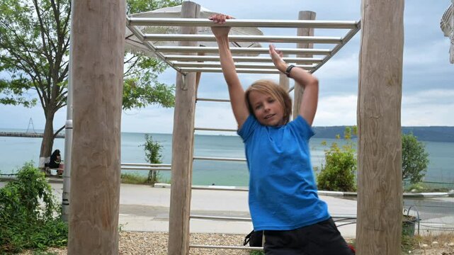 A boy in a blue shirt plays on a monkey bar near the seaside, enjoying an outdoor adventure. The scenic ocean backdrop adds a peaceful yet active feel to the moment