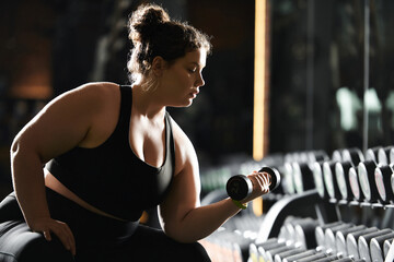 Fototapeta premium A young plus-size woman focuses on her workout, lifting a dumbbell with determination in a vibrant gym.
