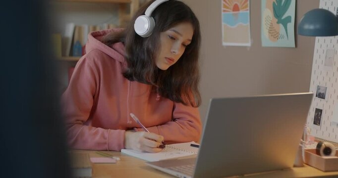 Schoolgirl doing homework with online tutor writing and looking at laptop screen listening to lecture with headphones at desk at home. Education and teenager concept.