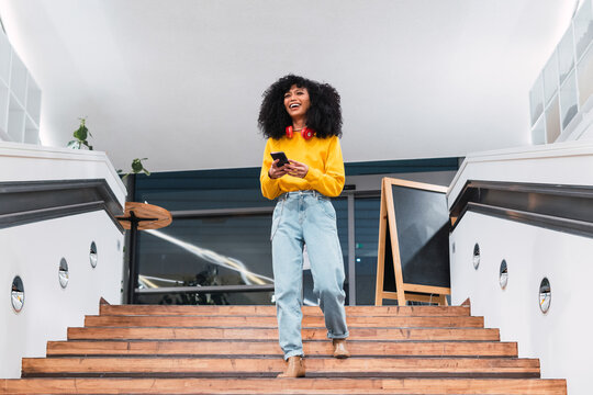 Photo of cheerful delighted African American woman types sms on modern cellphone device, enjoys good internet connection, dressed in yellow jumper
