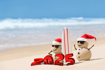 Two sandy snowmen on a beautiful beach, wearing sunglasses and with a surfboard. Large 3D numbers for the new year stand in front of them in the sand.