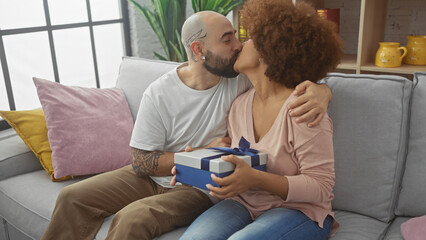 Interracial couple kisses while exchanging gifts, showcasing love and affection in a cozy home environment.