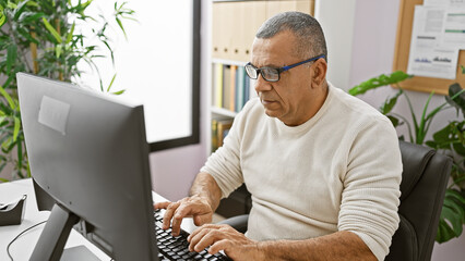 Hispanic man typing in a modern office surrounded by plants, exuding professionalism and experience.