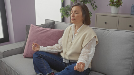 Mature woman meditates peacefully in a well-lit living room, embodying tranquility and mindfulness.