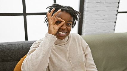 Smiling young black woman with dreadlocks making an okay sign indoors on a sofa, portraying a relaxed and playful mood.