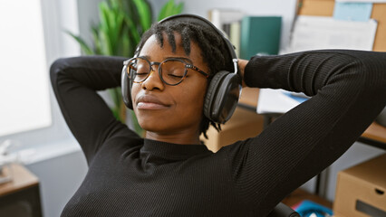 Fototapeta premium A relaxed african american woman with dreadlocks enjoys music on headphones in a modern office setting.