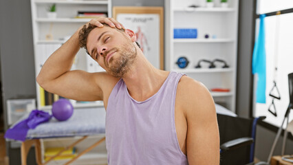 A young hispanic man with a beard stretching his neck in a rehabilitation clinic setting, conveying health and wellness.