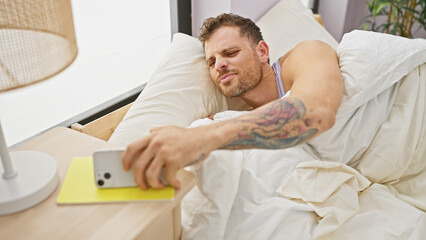 Obraz premium A tattooed young man with a beard reaches for his smartphone in the bedroom upon waking.