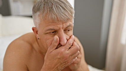 Obraz premium A pensive middle-aged man with grey hair sitting shirtless in a bedroom, touching his face with a thoughtful expression.