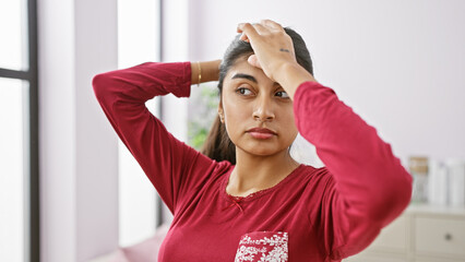 A young indian woman with long hair wearing a red shirt poses casually in a modern bedroom setting.