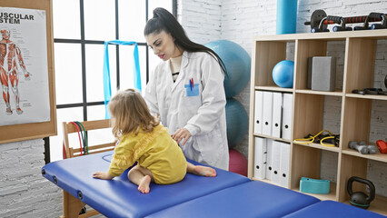 Pediatrician woman examining little girl in a rehab clinic room with physiotherapy equipment.