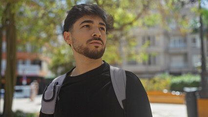 Handsome young man with a beard wearing a casual outfit, looking away thoughtfully in a vibrant city park during the daytime.