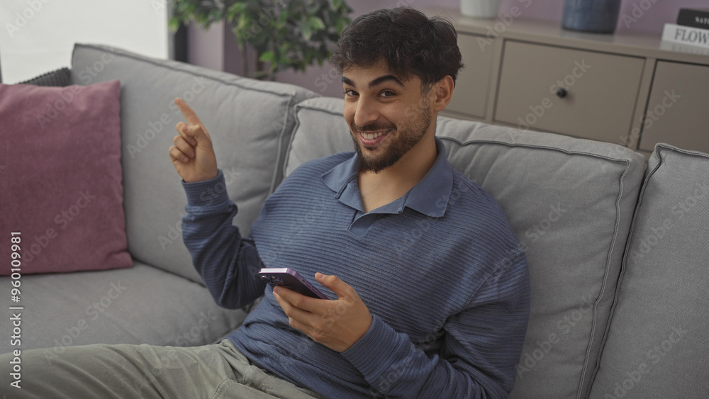 Wall mural a smiling young man with a beard, using a smartphone, gestures while lounging on a sofa indoors.