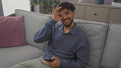 A cheerful young man makes an ok gesture over his eye while holding a smartphone in a modern living room.