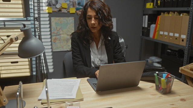 Mature hispanic woman working on laptop in a detective's office filled with files and a map.