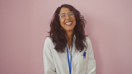 A joyful hispanic woman laughs heartily in a white lab coat against a pink background.