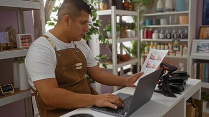 Young man in apron working on laptop and analyzing documents in a bakery shop with shelves of...