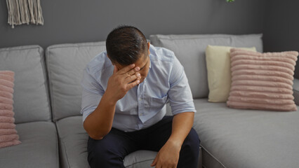 Hispanic man feeling stressed sitting on a sofa in a living room with a gray interior and decorative pillows in an apartment.