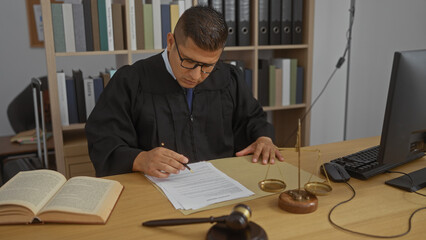 Young hispanic man working as a judge in an office, signing legal documents at his desk with a computer, law books, and a gavel