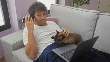 Hispanic man with a cat listens to a voice message in a living room while using a laptop on a couch indoors.