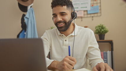 A cheerful young man with a headset engaged in a remote conversation at his modern apartment, pen in hand, working.