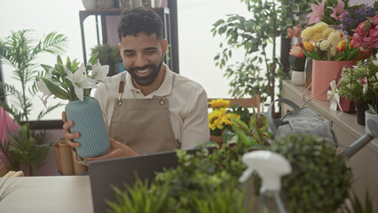 A smiling young man with a beard conducting a video call in a flower shop surrounded by colorful...