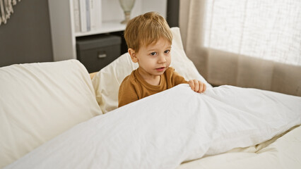 Fototapeta premium A pensive blond toddler sits on a bed in a well-lit, cozy bedroom, exuding an air of innocence and childhood.