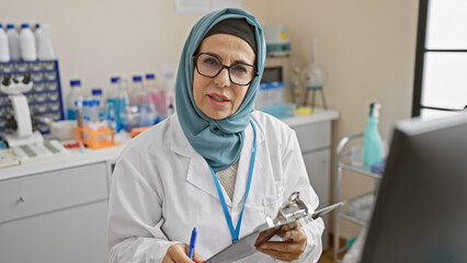 A mature woman in hijab and lab coat taking notes in a clinic laboratory setting.