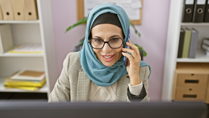 A professional woman in hijab smiling while talking on the phone in a modern office setting.
