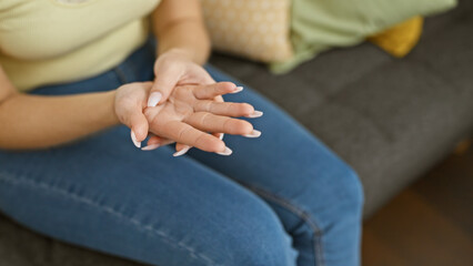 A young woman examines her hand, suggesting pain or injury, while sitting indoor in a comfortably furnished living room.