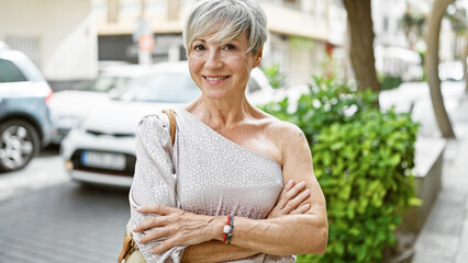Elegant mature hispanic woman with grey short hair, crossing her arms, poses on a sunny urban street.