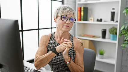 Mature woman with glasses and short grey hair contemplates in modern office setting.