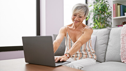 Mature woman using laptop cushioned by sofa in a cozy room, exuding comfort and casual lifestyle.